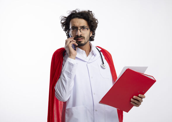 持有无知的年轻白种人超级英雄 戴着眼镜 穿着医生制服 披着红色斗篷 脖子上戴着听诊器 一边打电话一边拿着文件夹和复印件文件年轻脖子