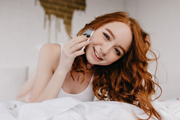 电话欣喜若狂的红发女孩躺在床上拿着手机浪漫的欧洲女模特拿着智能手机在卧室摆姿势智能手机房间年轻