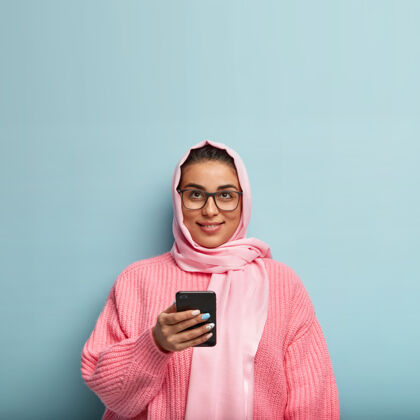 面纱一个美丽的梦幻女人用手机在社交网络上发送信息的孤立镜头 向上聚焦 思考信息的内容 站在蓝色的墙上思考阿拉伯小玩意