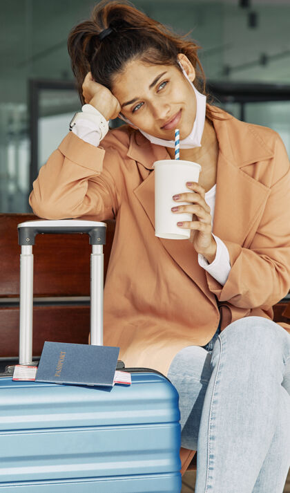通勤在大流行期间 一个女人在机场的行李上休息 喝咖啡流行病医疗口罩行李