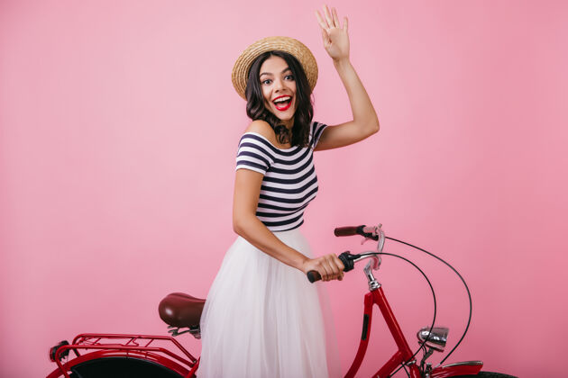 年轻戴草帽的快乐女人在自行车上摆出惊奇的微笑室内拍摄的迷人的黑发女孩表达快乐的情绪微笑快乐时尚