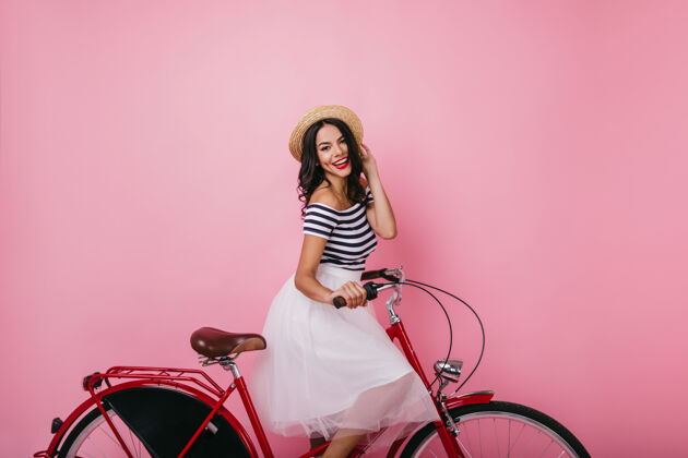 欢快兴奋的拉丁女孩坐在自行车上 带着愉快的微笑穿着华丽裙子的时髦女士的画像卷发女人帽子