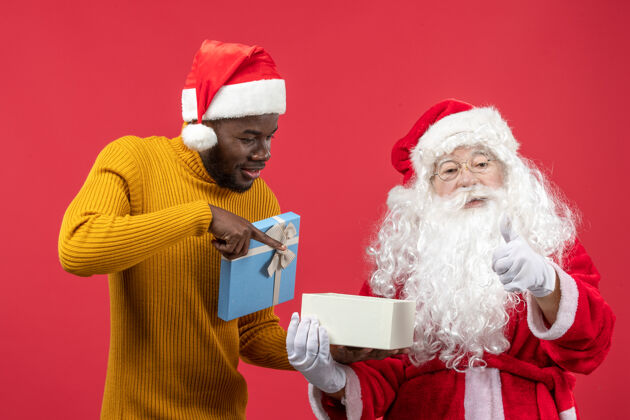 礼物圣诞老人和年轻人在红墙上打开礼物的正面视图快乐圣诞快乐帽子