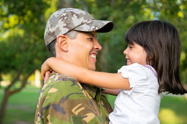 伪装身着迷彩服的快乐爸爸抱着小女儿 在军事任务归来后在户外拥抱女孩特写镜头家庭团聚或回家的概念军人力量公园