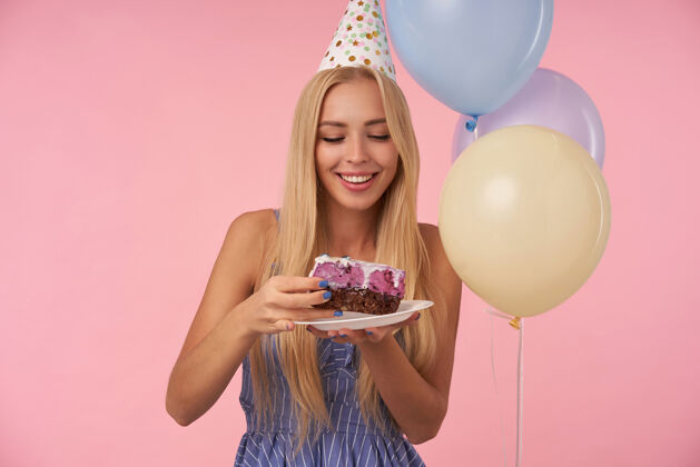 吹穿着节日服装 戴着圆锥形帽子 站在粉色背景和五颜六色的氦气球上 准备吃节日蛋糕的快乐的年轻金发女郎 展示了她愉快的心情圆锥形饮食享受