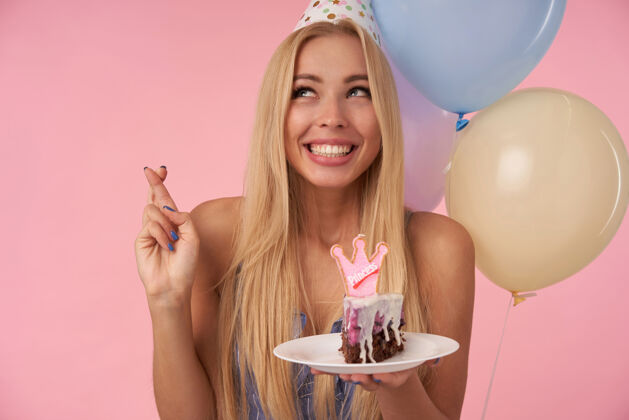 迷人快乐的年轻长发女性 金色长发 在生日许愿时交叉手指 拿着一块蛋糕和蜡烛在五颜六色的气球和粉红色的背景上手指气球吹