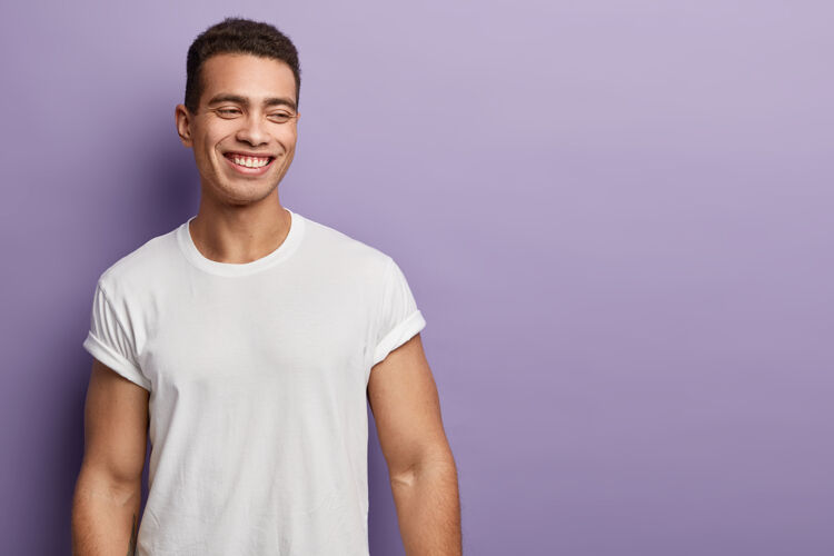 设计帅气肌肉男的室内镜头 表情积极 穿着休闲的白色t恤 靠着紫色的墙壁 自由的空间放在一边人和情感积极男人友好