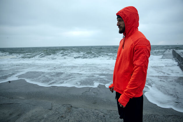 海滨在灰色的清晨 身穿暖橙色连帽衫和黑色运动裤的年轻黑发胡须运动员站在海边 若有所思地向前看户外运动员运动型