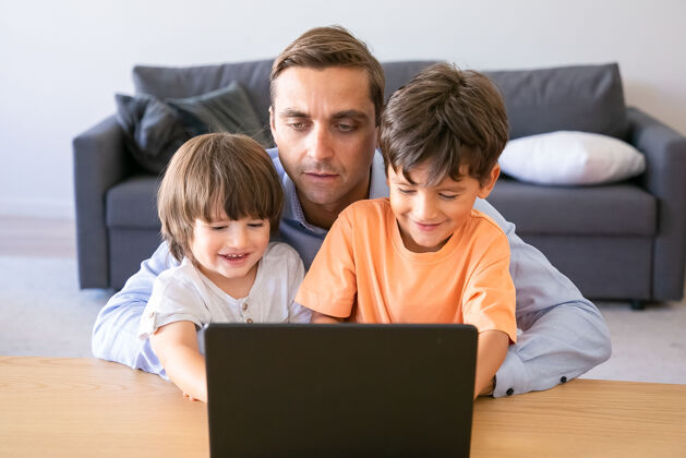玩爸爸抱着儿子在笔记本电脑上看电影的正面图两个可爱的小男孩和爸爸坐在桌子旁 看着屏幕 微笑着父爱 童年和数字技术概念男孩爸爸孩子