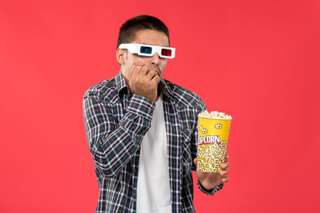 成人正面图：戴着d型太阳镜的年轻男子拿着爆米花在浅红色的墙上看男性电影帅哥浅红色电影院