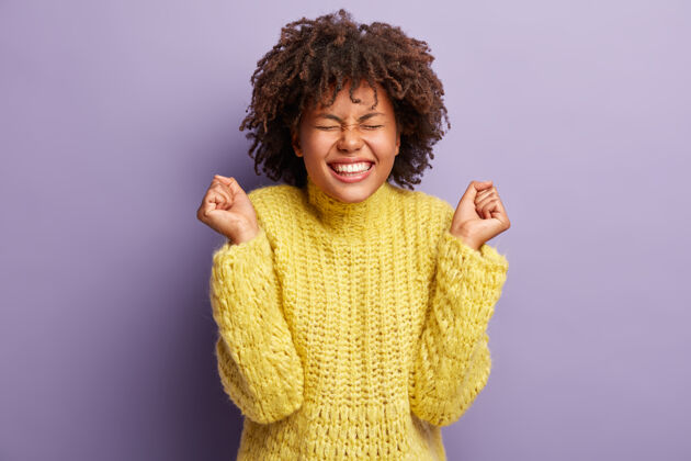 成人喜出望外的黑人妇女庆祝成功的镜头 有成功 穿着黄色的套头衫 露出白色的牙齿 有牙齿的微笑 对着紫色的墙壁庆祝的概念幸福拳头展示