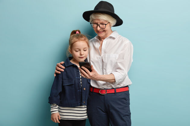 领养老金者家庭 时代和科技概念快乐时尚的祖母手持智能手机 拥抱女孙 一起看视频 一起在家里玩得开心 在蓝色的墙上摆姿势小智能手机小玩意