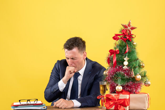 会议忧心忡忡的男人闭上眼睛坐在圣诞树旁的桌子上 用黄色的礼物忧虑的男人坐着眼睛