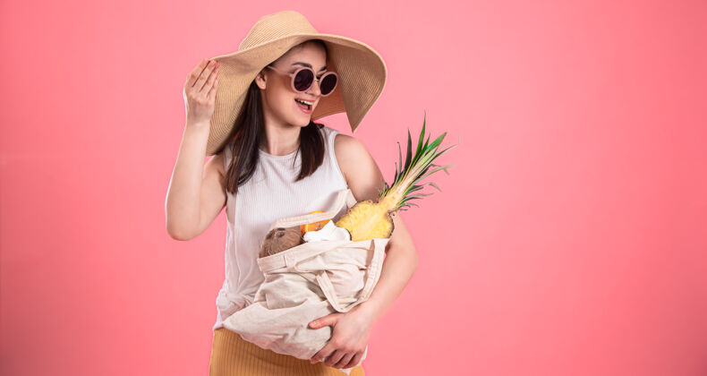 夏天戴着大帽子和太阳镜的时髦女孩微笑着 手里拿着一个装有异国水果的环保袋包帽子女孩