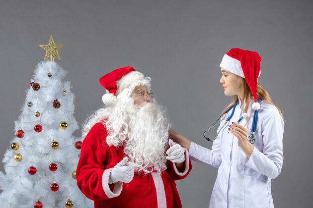 十二月圣诞老人和女医生的前视图 女医生拿着测温仪在灰色的墙上举行温度节日
