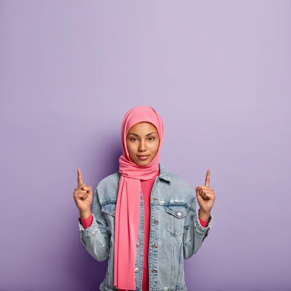 室内自信自信的女人 表情沉稳 指向上方的自由空间 头戴头巾 吸引你的注意力 隔离在紫色的墙壁上广告和种族概念摆姿势宗教传统