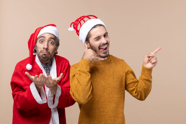 男人正面图两个戴着圣诞帽的朋友一个在米色背景上打电话给我前面男性圣诞老人