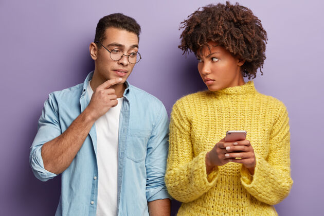 手机多种族的夫妇在手机上得到消息愤怒的女人要求男友不要偷看手机 看着不满意 穿着黄色的套头衫好奇的戴眼镜的白人男人有兴趣打字短信黑暗