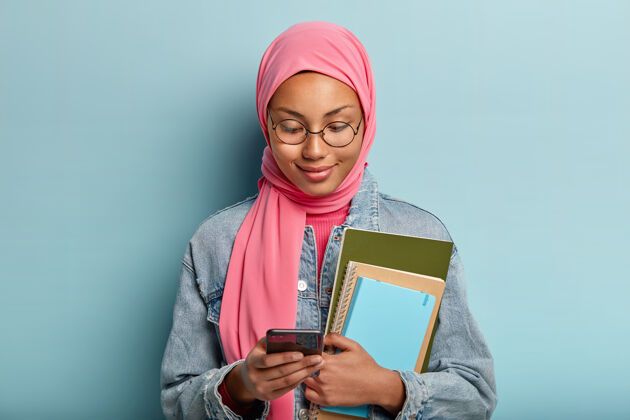 打字迷人的阿拉伯混血女子头像专注于手机 在手机上处理文件 喜欢新的应用程序 手持螺旋笔记本 身着牛仔夹克和粉色头巾 根据宗教信仰伊斯兰教无线手机
