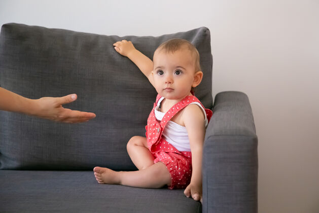 新生儿有趣的小宝宝坐在灰色沙发上 看着一个不认识的人有人向穿着红色短裙的可爱小女孩伸出手来家庭 童年和家的概念生活小地毯