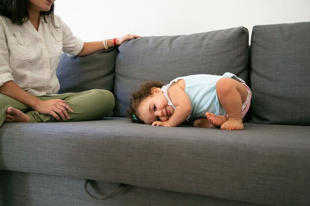 成人穿着浅蓝色连衣裙的可爱有趣的女婴躺在妈妈旁边的灰色沙发上不规则镜头父母和童年的概念父母衣服母亲