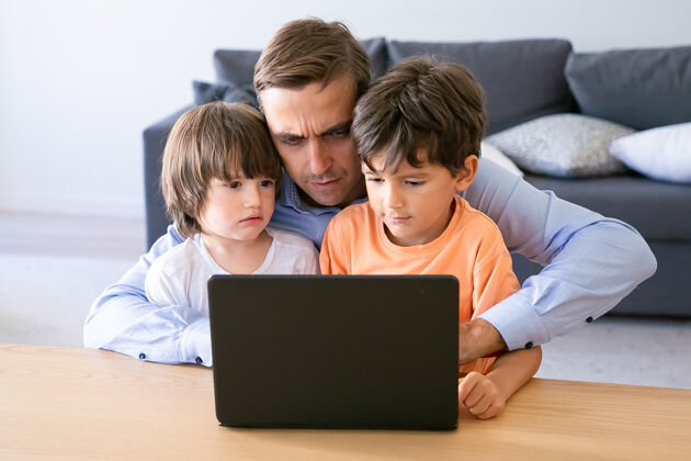 使用严肃的爸爸在笔记本电脑上工作 拥抱儿子专注的白人爸爸在家里使用笔记本电脑两个可爱的男孩坐在他的膝盖上父亲 童年和数字技术的概念儿子严肃可爱