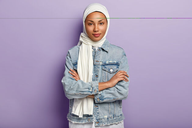 自信迷人的东方女人头戴白色头巾 以保护自己的尊严和权力 有特殊的着装要求 双手交叉 看起来谦虚 在紫色的墙壁上摆姿势伊斯兰规则个人外表伊斯兰教