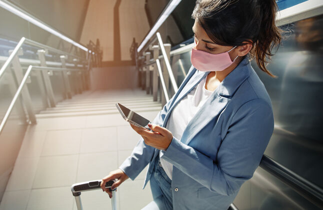 智能手机大流行期间在机场使用智能手机的戴着医用面罩和行李的高角度女性Covid19冠状病毒楼梯