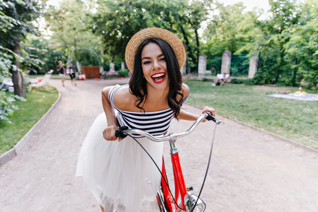 人戴着夏日帽子的快乐棕褐色女孩在公园里表达快乐穿着裙子的可爱黑发女人在大自然中与自行车合影的户外镜头骑欢笑魅力