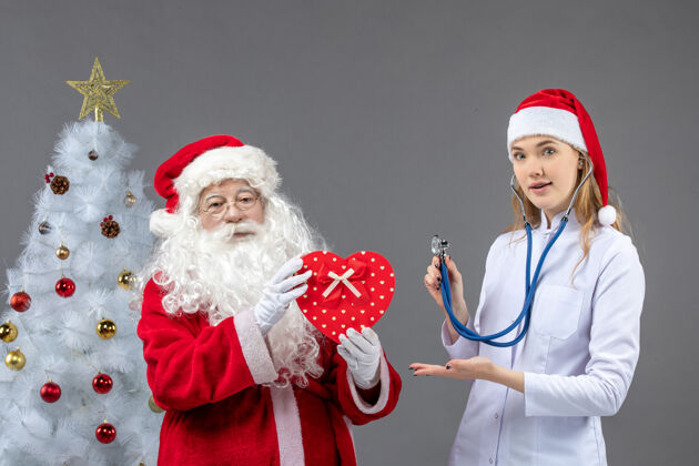 快乐圣诞老人和女医生在灰色墙上用听诊器观察的正视图正面医药听诊器