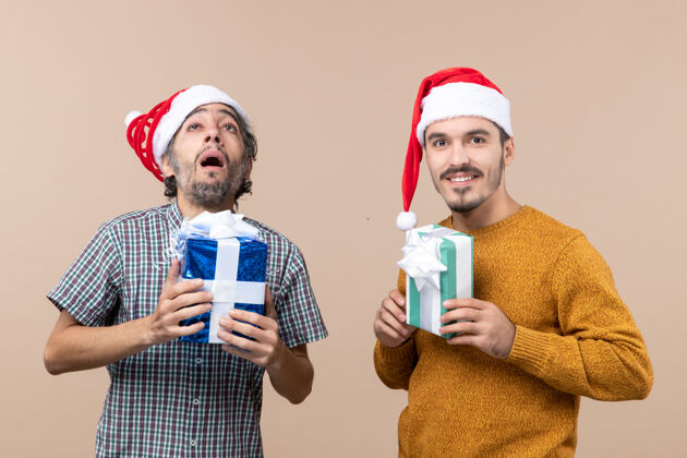 戴着正面图两个男人一个沮丧 一个快乐 戴着圣诞帽 拿着圣诞礼物 背景是米色的伙计们两个沮丧