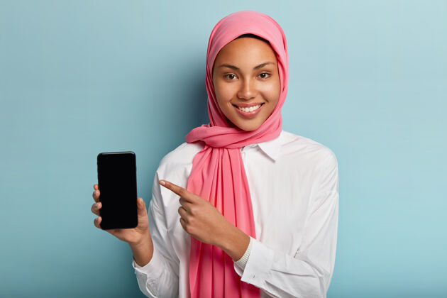 高兴女士手持智能手机 显示空白屏幕插入文本或您的信息 戴着粉色头巾和白色衬衫屏幕正面女士