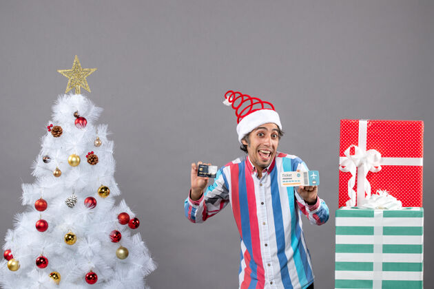 快乐的年轻人正面图快乐的年轻人拿着卡片和旅行票围着圣诞树和礼物手持人树