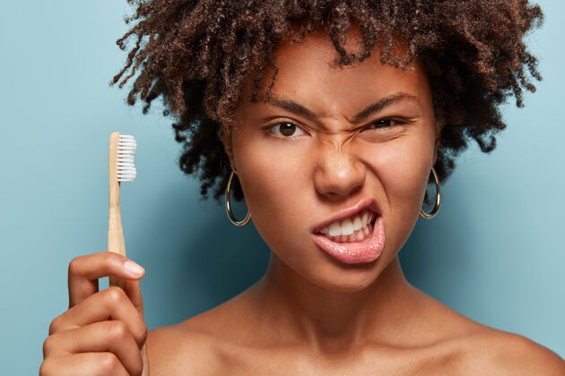 新鲜不高兴的非洲女人皱着眉头 咬紧牙关 注意口腔卫生 卷发 拿着牙刷 光着肩膀 在蓝色的墙上摆姿势清洁紧握个人
