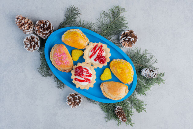 小吃蓝色盘子上放着小面包 纸杯蛋糕和果冻糖果 大理石表面装饰着松叶和圆锥体甜点糖美味
