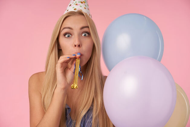 发型迷人的金发女郎 随意的发型 头戴蓝色圆锥形帽 吹派对喇叭 用五彩气球庆祝生日 在派对中度过快乐的时光庆祝吹正面