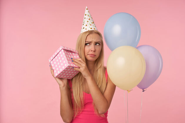 室内迷惑不解的长发金发女士 随意的发型 手里拿着礼物盒 试图找出里面是什么 用五颜六色的气球庆祝生日 在粉色背景上摆姿势发型圆锥体盒子