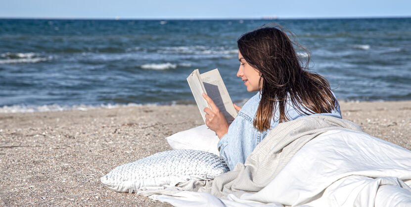 书一位年轻的黑发女子躺在海边 身上裹着毯子 正在看书海滩上的舒适气氛 夏日概念黑发舒适夏天