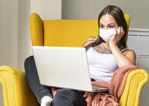 医疗口罩大流行期间 戴着医用面罩的妇女坐在扶手椅上操作笔记本电脑的正面图隐居距离妇女