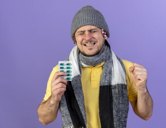 拳头疼痛的年轻金发病斯拉夫男子穿着和放在冬季帽子和围巾包药丸持有包药丸保持在紫色的墙上拳头包药片医学男人帽子