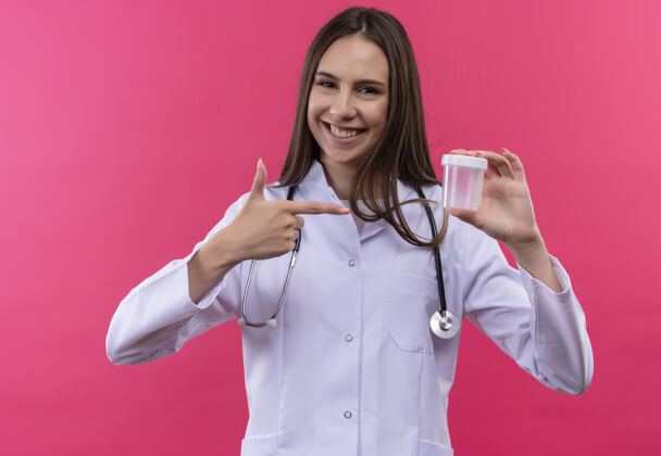 手微笑的年轻医生女孩穿着听诊器医用长袍指着她手上的空罐 背景是孤立的粉色长袍穿听诊器