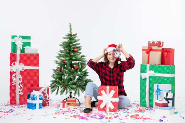 快乐前视图年轻女子坐在圣诞礼物周围拿着白色墙上的时钟时钟庆祝十二月