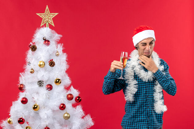 玻璃圣诞心情酸溜溜的脸年轻人戴着圣诞老人的帽子 穿着蓝色的衬衫 在圣诞树旁拿着一杯酒人夏娃庆祝