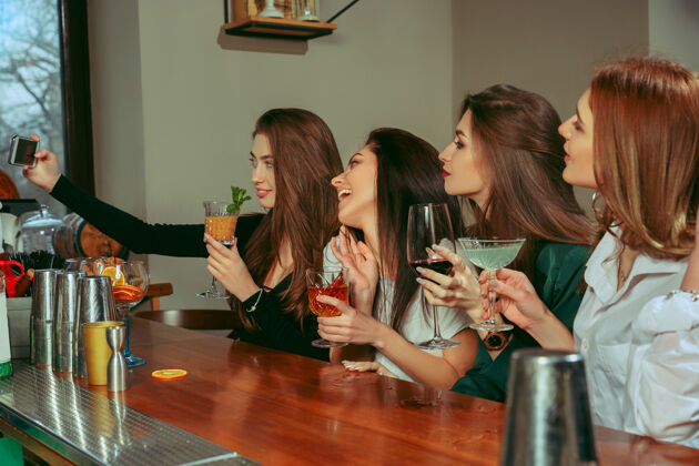 团体女朋友在酒吧喝酒她们坐在一张摆着鸡尾酒的木桌旁她们穿着休闲服朋友酒精年轻
