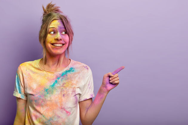 胡里节快乐快乐的女性玩弄胡里色彩 在节日里玩得很开心 用食指指着 广告复制空间 喜欢鲜艳的颜色洒在脸上和t恤上 在紫色的墙上做手势积极过度运动牙齿