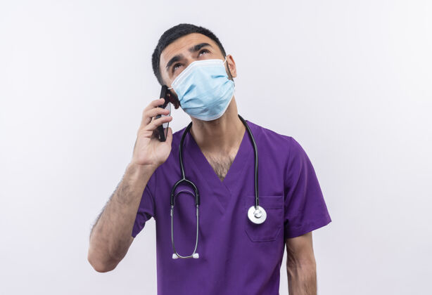 看看着身穿紫色手术服和听诊器医用面罩的年轻男医生在孤立的白色背景下讲电话衣服年轻紫色