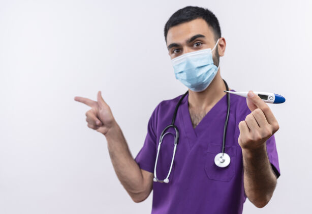 医疗年轻的男医生穿着紫色手术服 戴着听诊器医用面罩 手持体温计 站在隔离的白色背景上外科医生侧面年轻人
