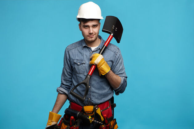 劳工年轻的挖掘机戴着手套 戴着白色头盔 工具带上铲子的照片防护挖掘牛仔裤
