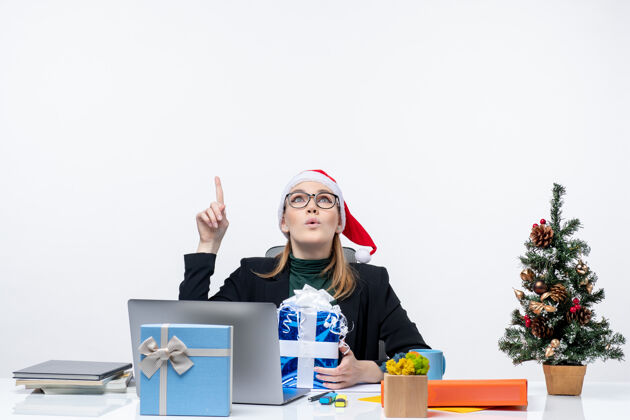 圣诞老人圣诞节的心情与惊讶的年轻女子戴着圣诞老人的帽子和眼镜坐在一张桌子上拿着礼物 并显示在白色背景以上圣诞老人坐着眼镜