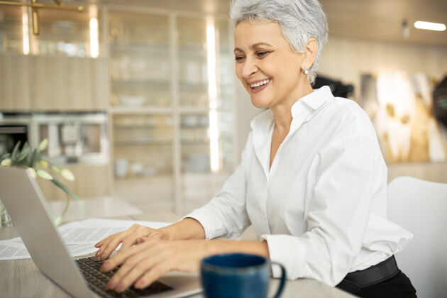 咖啡侧视图：快乐的中年女商人 灰色短发 在时尚的办公室里用笔记本电脑工作 手放在键盘上 打字 分享好消息女商人工作电子邮件
